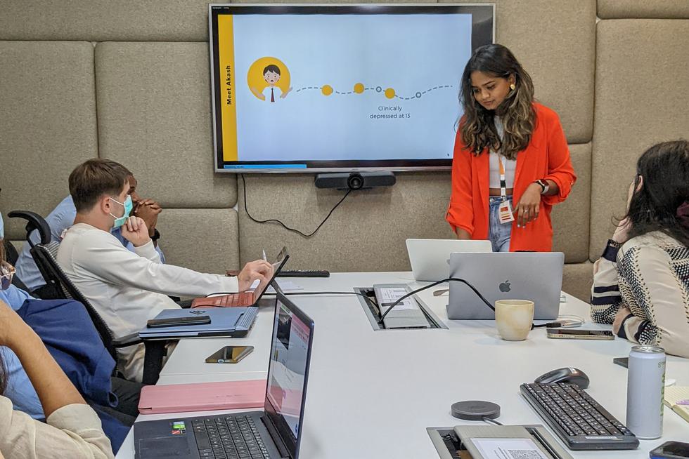 Kavindya Thennakoon leads a meeting. She stands at the front while others sit around a table. A screen is on the wall.