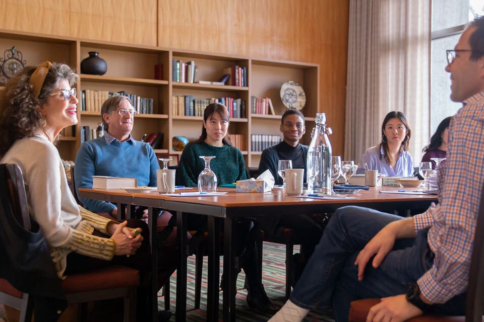A group of students and a guest speaker sit around a table, having tea.