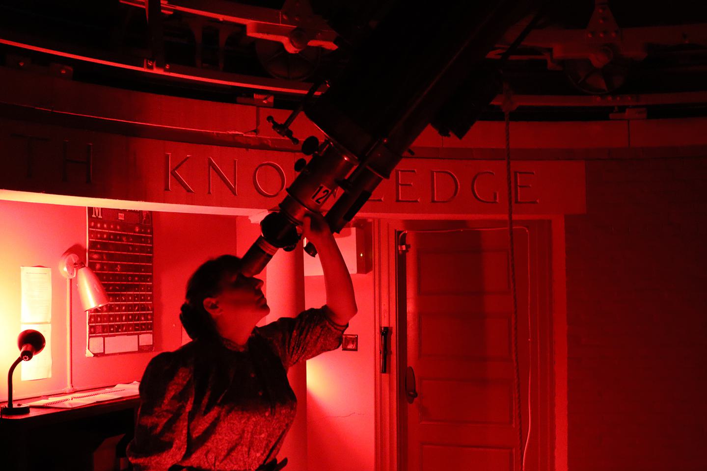 A woman looks through a telescope inside Whitin Observatory. She is bathed in red light.
