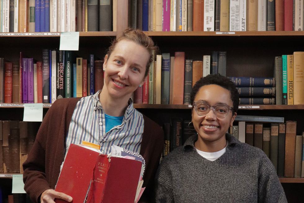 Corinne Gartner, associate professor of philosophy, and Adele Watkins, assistant professor of philosophy pose in front of their bookshelves filled with philosophy books. They each hold a book.