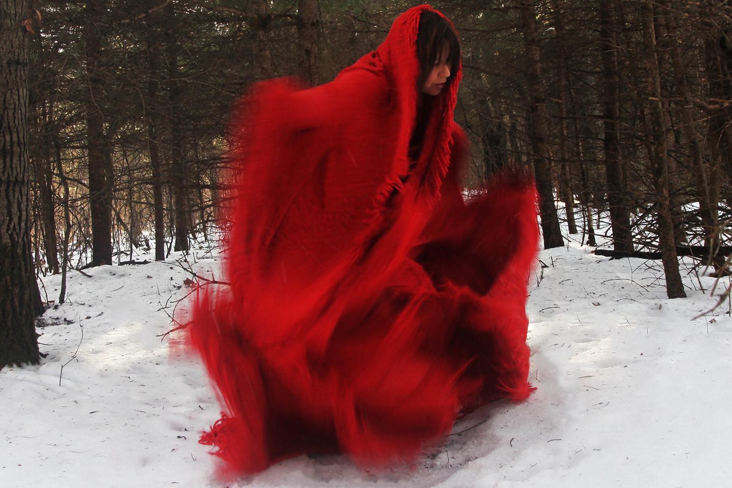 A photo shows a woman in movement, wearing a red blanket-like dress and surrounded by trees with snow on the ground.