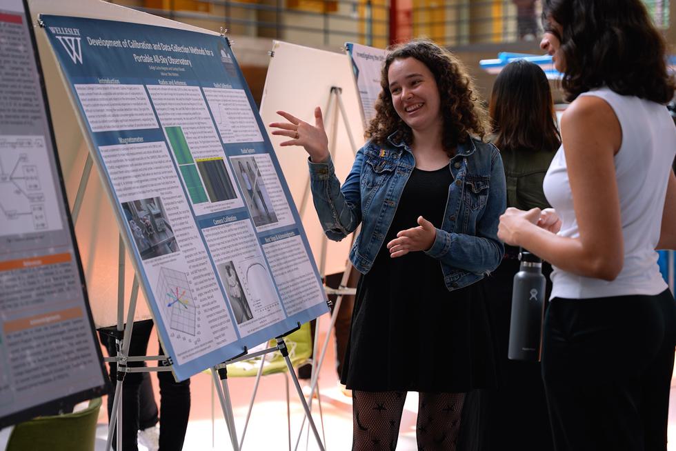 Student gestures toward her science research poster, talking with a visitor.