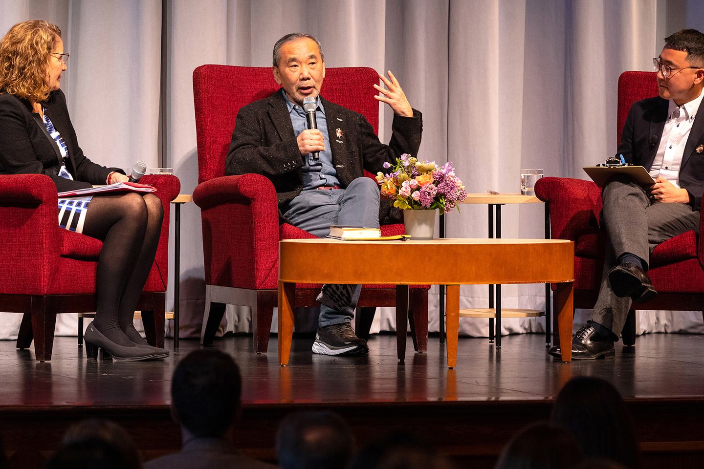 Haruki Murakami seated on stage and holding a microphone to answer a question.