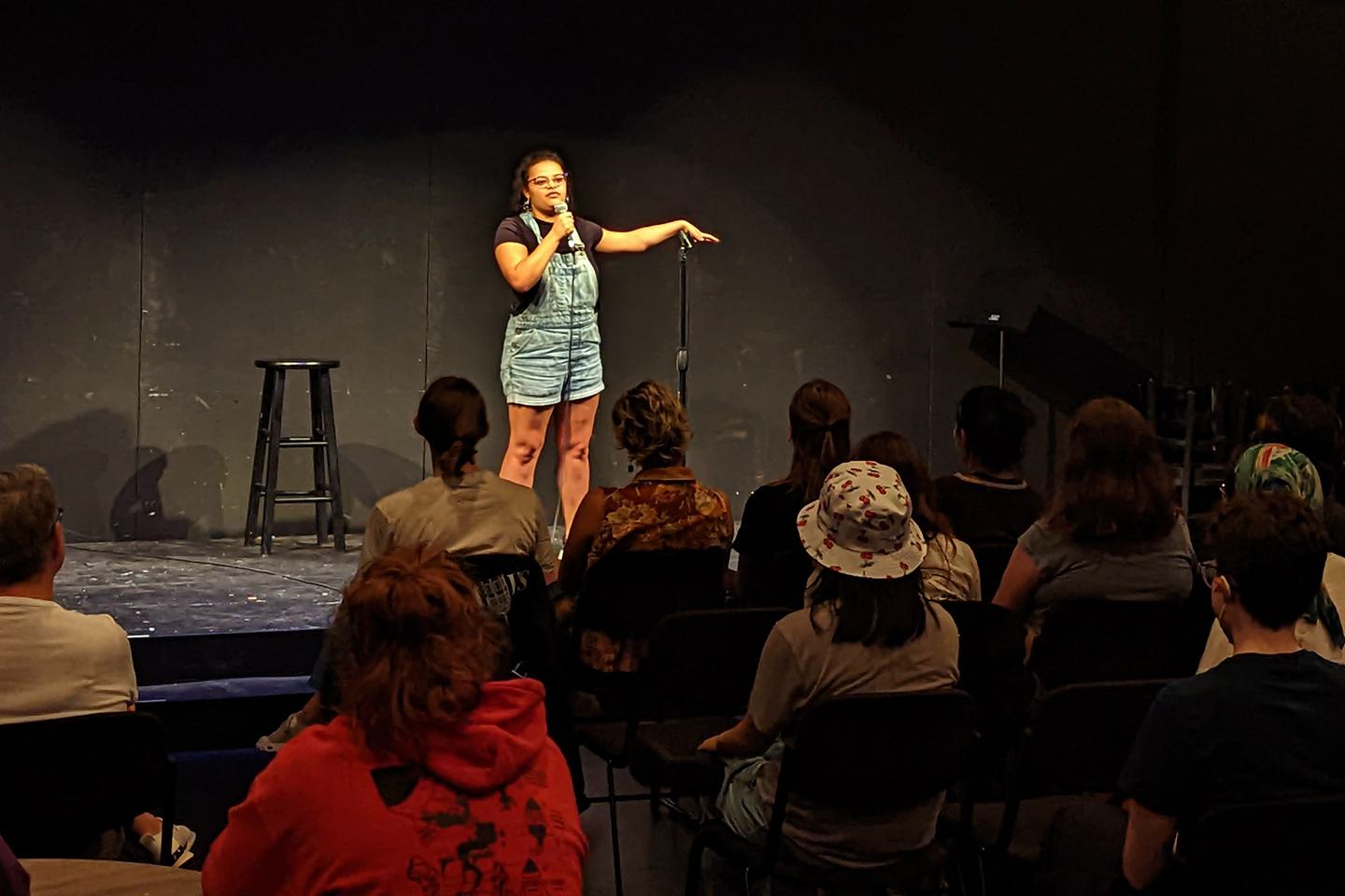 A student stands in the spotlight on stage, performing stand-up comedy for an audience.