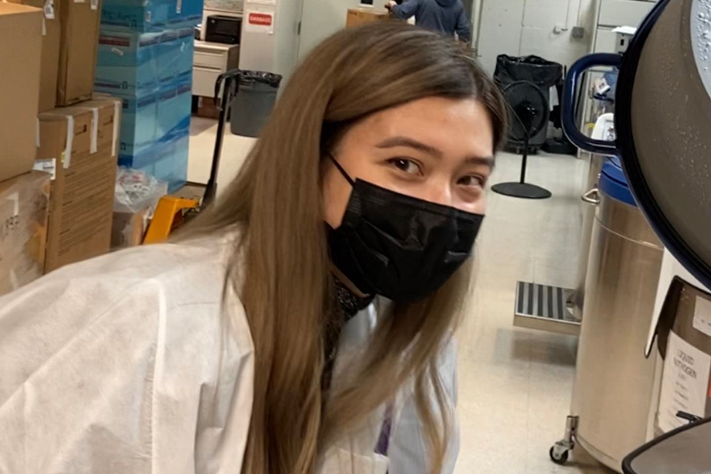 Kathryn Cross in a lab coat and black face mask looks at camera.