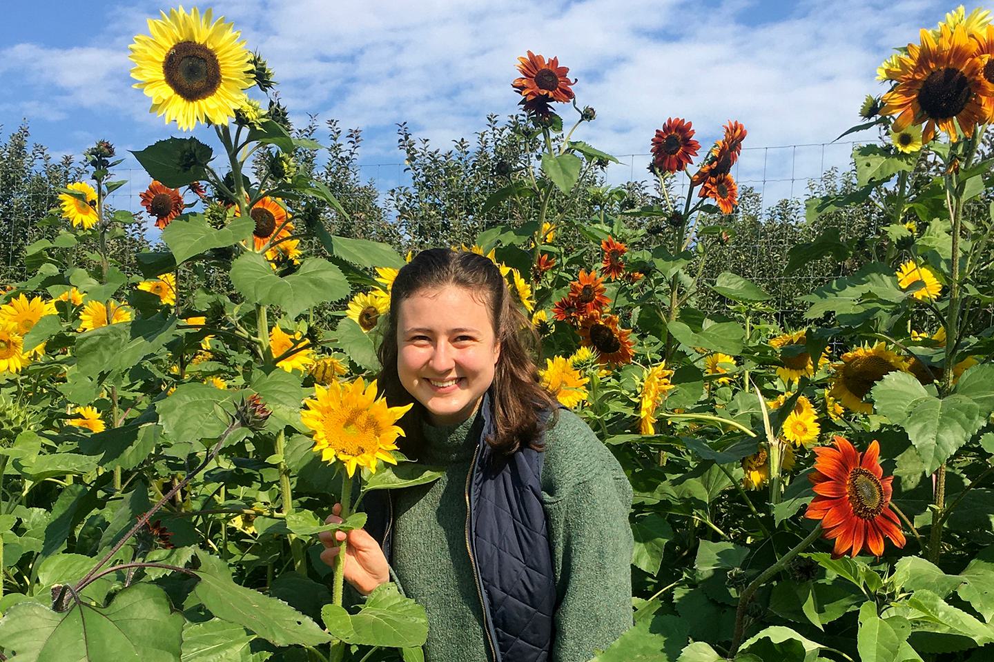Dominique Mickiewicz stands in a garden of sunflowers and smiles at the camera.