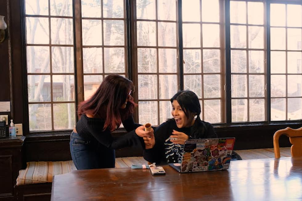 A student passes off a paper baton to another student, who expresses great excitement with her face.