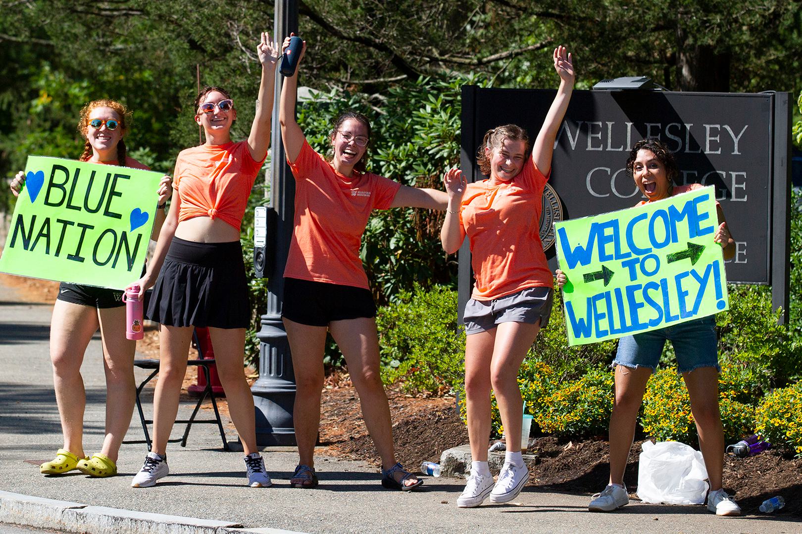 Five students stand at the entrance of Wellesley, waving welcome signs.