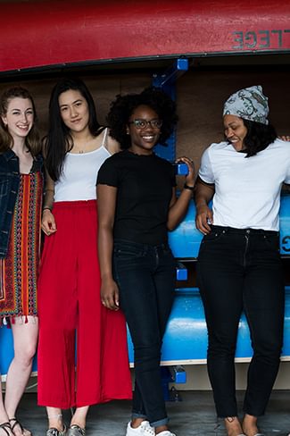 Five Wellesley College students pose for a photo in front of blue canoes in a boat house