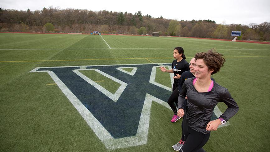 Wellesley student athletes run on a sports field with the Wellesley logo